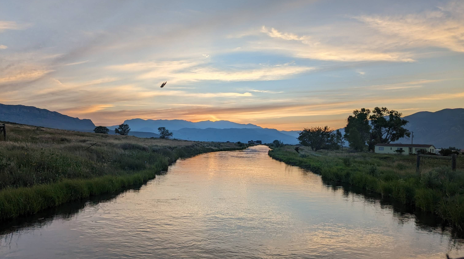 A peaceful river flows between grassy banks at sunset, with mountains near charming tourist towns in the distance, colorful clouds overhead, and a bird flying above the water. A few trees and a small house are visible on the right side.