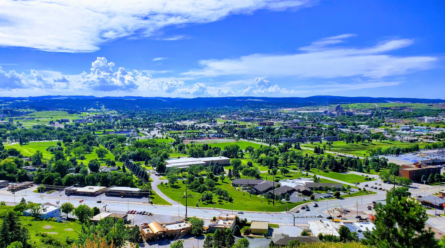 A scenic view of a city resembling charming tourist towns, with lush green trees, scattered buildings, and roads under a bright blue sky with white clouds and distant hills in the background.