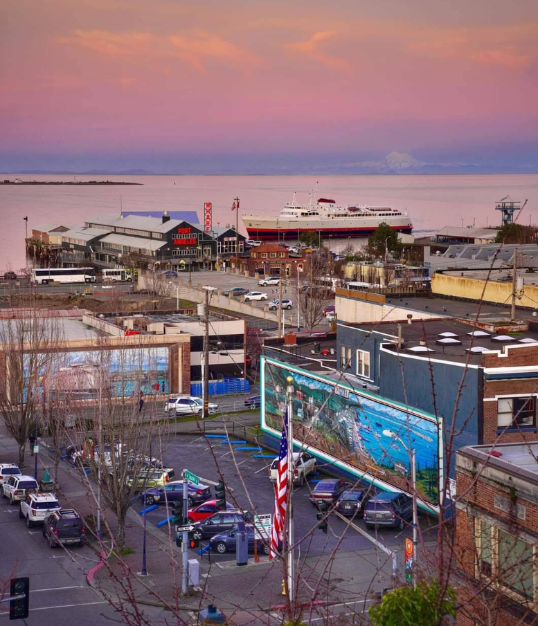 A coastal tourist town at sunset with a ferry at the dock, colorful sky, parked cars, American flag, and buildings adorned with murals and billboards in the foreground.