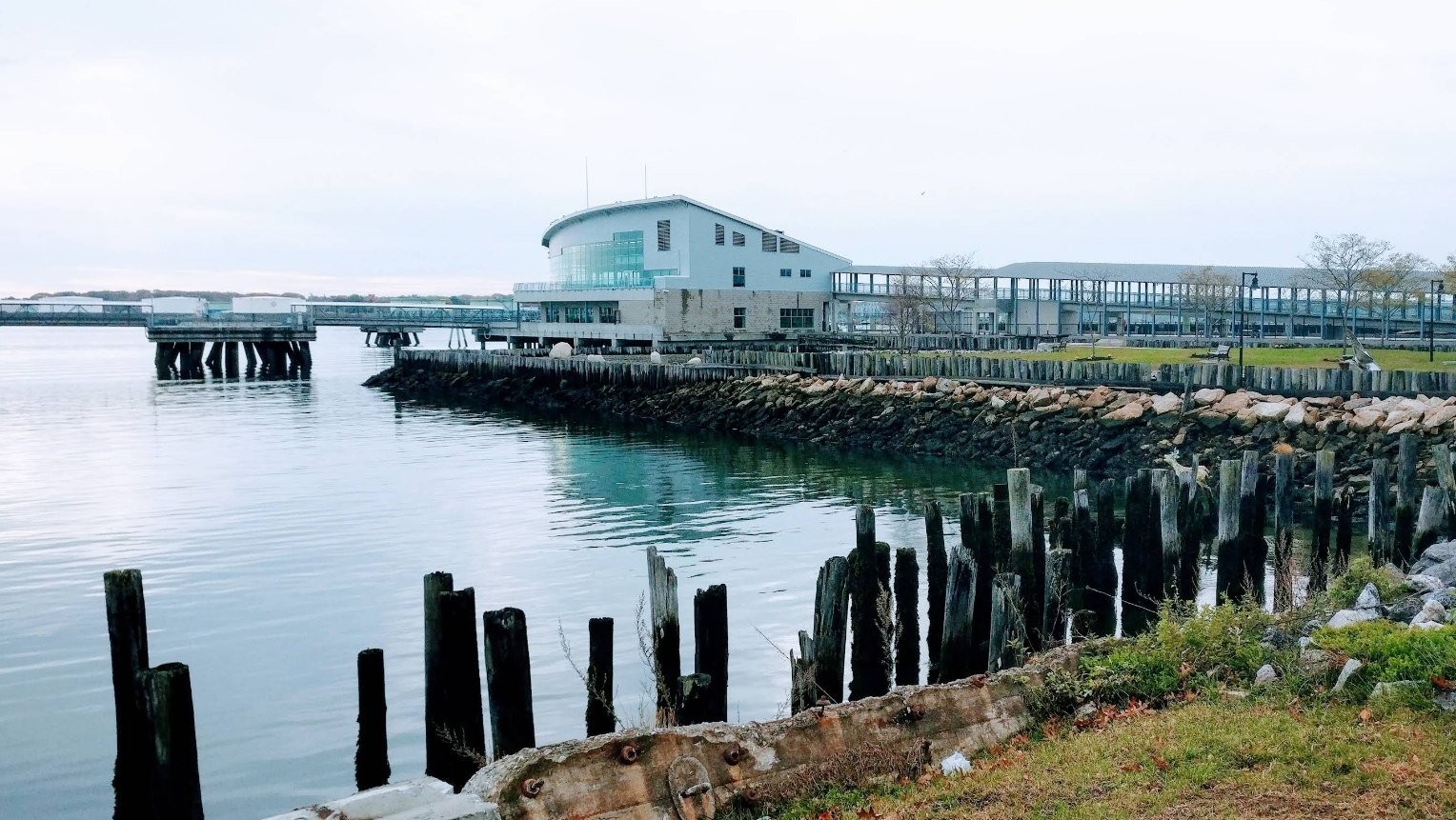 A modern building with large windows sits by the water in one of the region's charming tourist towns, surrounded by a rocky shoreline, wooden pier posts, and grassy area. The overcast sky and calm water reflect the peaceful setting.