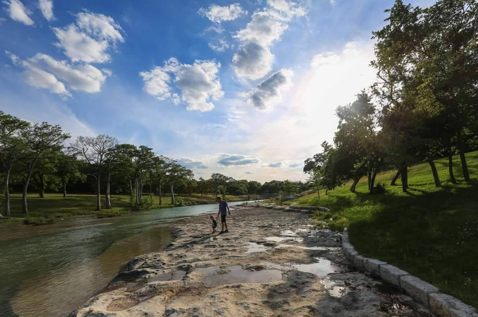 A person walks along a rocky riverside under a bright, partly cloudy sky. Trees line both sides of the river, common in scenic tourist towns, while sunlight casts shadows on the grass and rocks.