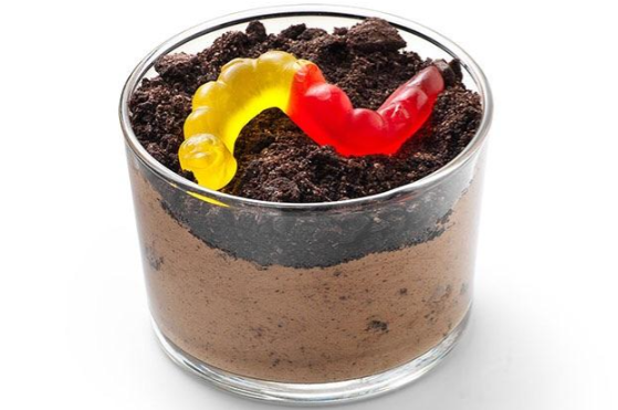 A glass cup filled with chocolate pudding, topped with crushed chocolate cookies and two gummy worms, resembling dirt and worms.