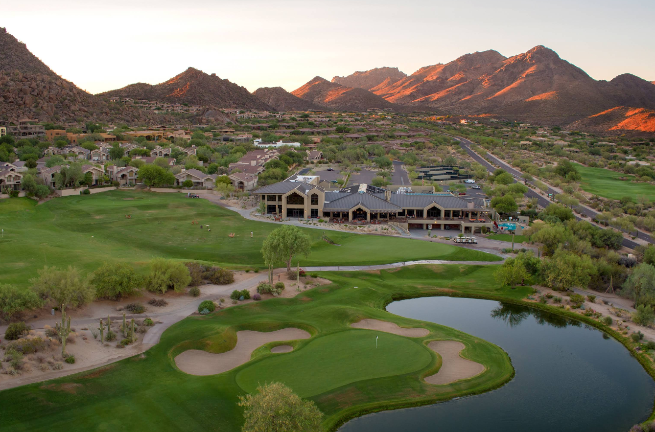 Aerial view of a golf course with green fairways, sand traps, a small pond, a clubhouse, scattered homes, and rugged mountains in the background at sunset—an inviting scene even as hotel prices have risen nearby.