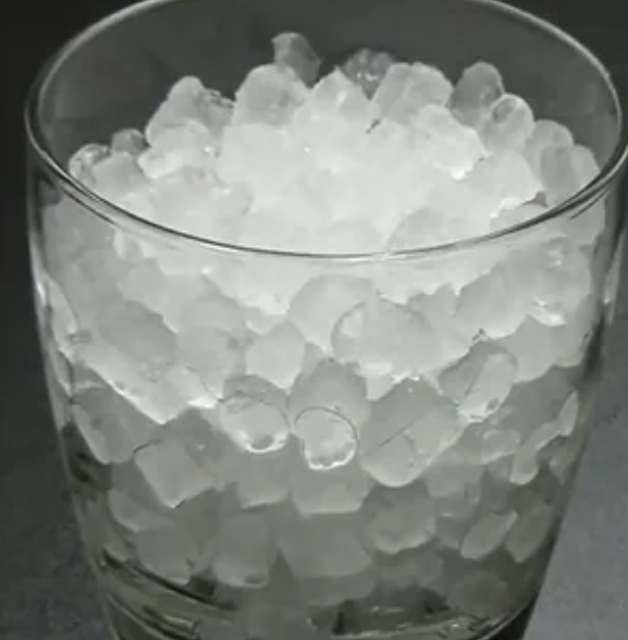 A clear glass filled to the brim with small, cylindrical ice cubes, sitting on a dark surface.