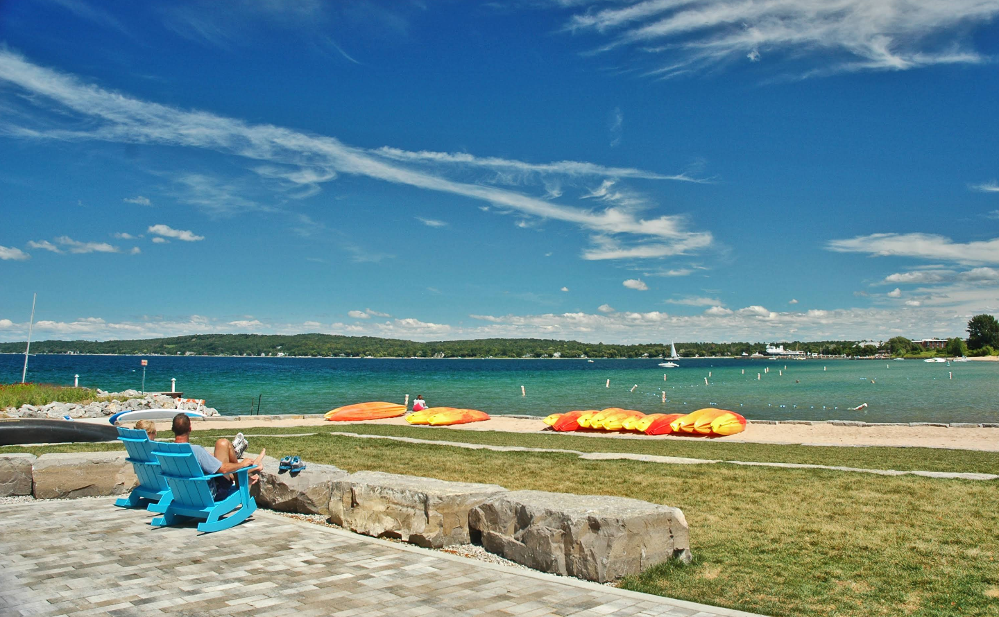 Two people relax in blue chairs facing a sandy beach and calm lake, typical of scenic lake towns. Brightly colored kayaks line the sand, with green hills and a partly cloudy sky in the background.