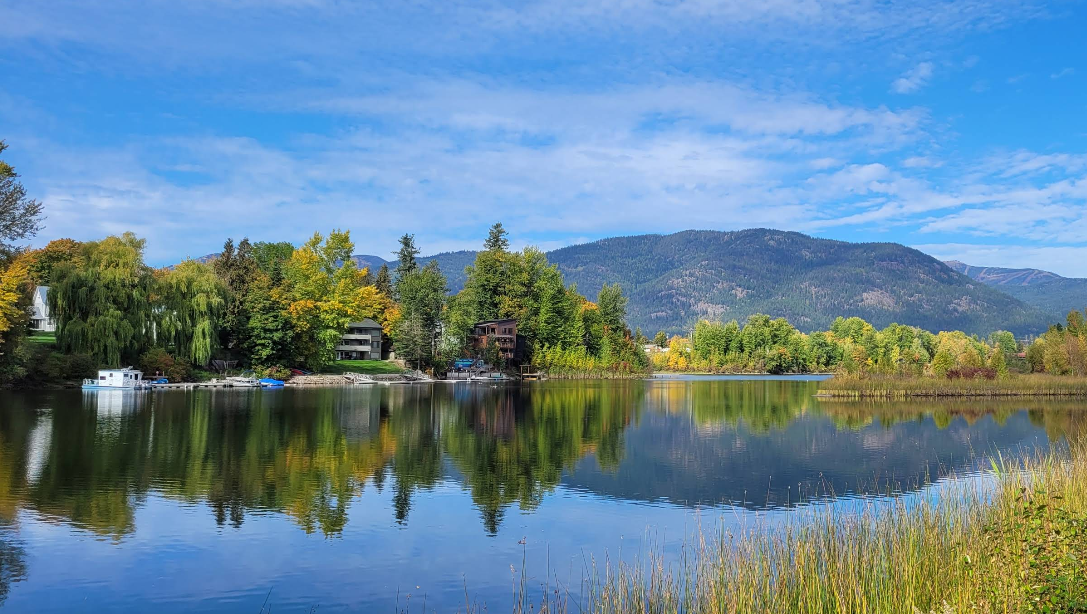 A calm lake in picturesque lake towns reflects trees with autumn foliage, houses, and a blue sky with scattered clouds. Forested hills and mountains rise in the background, while tall grass lines the water’s edge in the foreground.