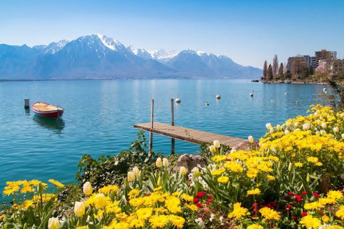 A serene lake town scene with clear blue water, a wooden dock, and a small boat. Snow-capped mountains rise in the background, while bright yellow flowers bloom in the foreground under a clear, sunny sky.