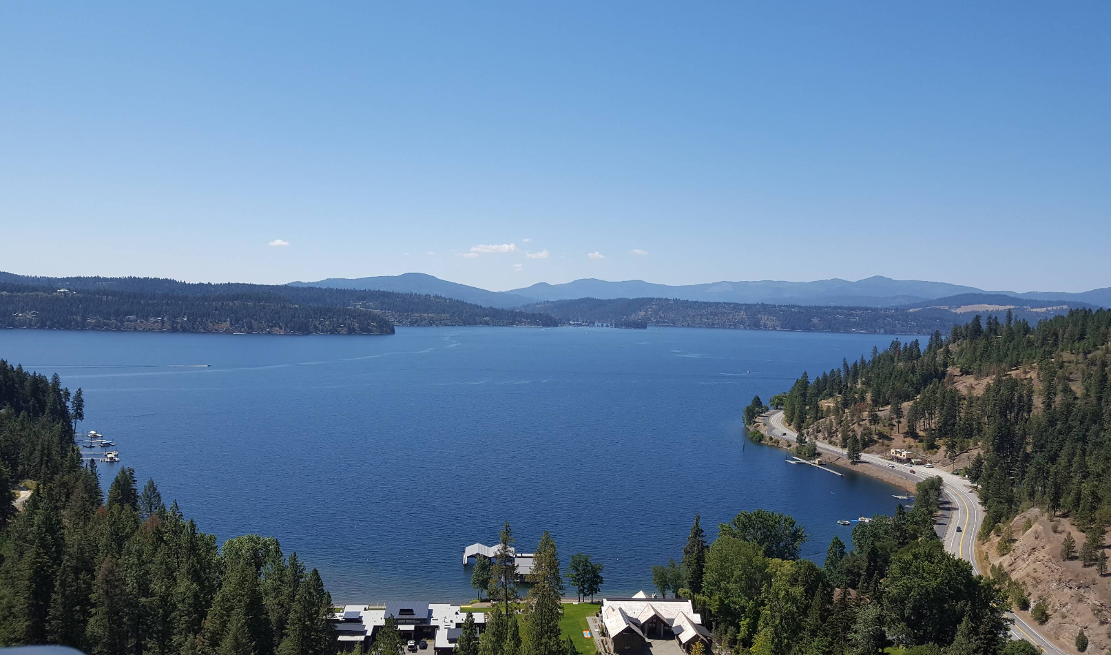 A large blue lake surrounded by green forested hills and mountains under a clear blue sky, with charming lake towns, a few buildings, and a winding road along the shoreline.