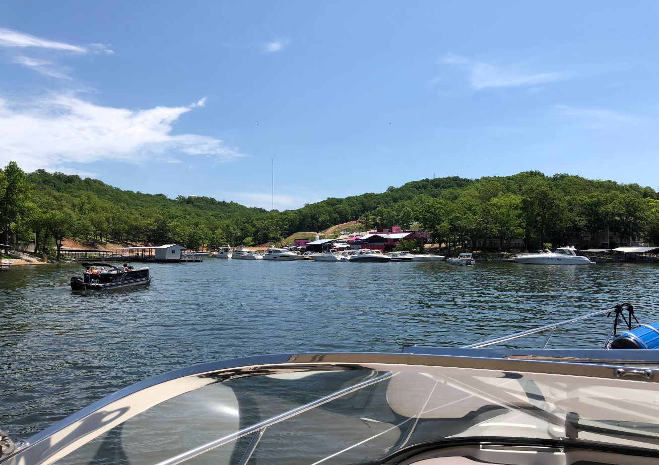 View from a boat on a calm lake in one of the charming lake towns, with several boats docked along the shore, green trees and hills in the background, and buildings near the water under a blue sky with scattered clouds.