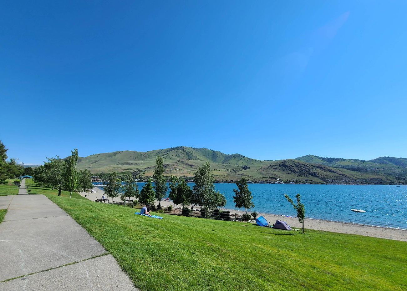 A paved walkway runs alongside a grassy area with scattered trees, leading to a sandy lakeshore. Hills and mountains rise in the background under a bright blue sky, capturing the charm of lake towns. Small boats and tents are visible near the water.
