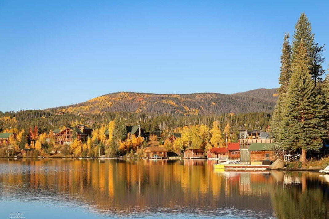 A calm lake in one of the charming lake towns reflects colorful autumn trees and wooden cabins, with forested hills and a bright blue sky in the background. The scene is peaceful and picturesque.