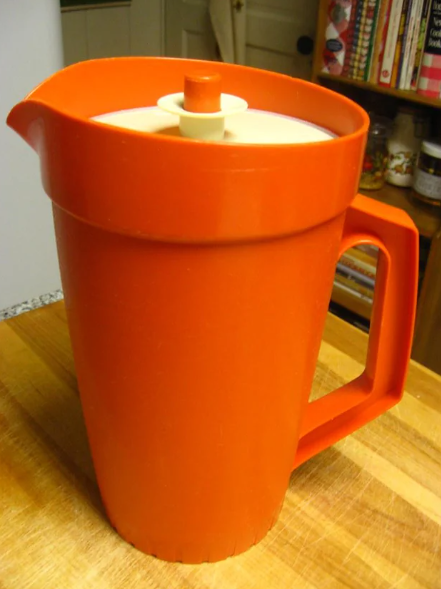 An orange plastic pitcher with a handle and a beige lid sits on a wooden countertop, with shelves and cookbooks visible in the background.