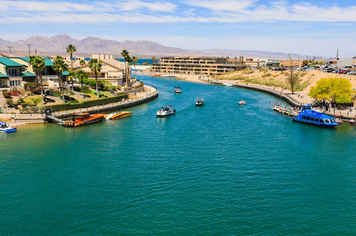 A bright, sunny day on a wide blue river in one of the charming lake towns, with several boats, surrounded by buildings, palm trees, and distant mountains under a partly cloudy sky.