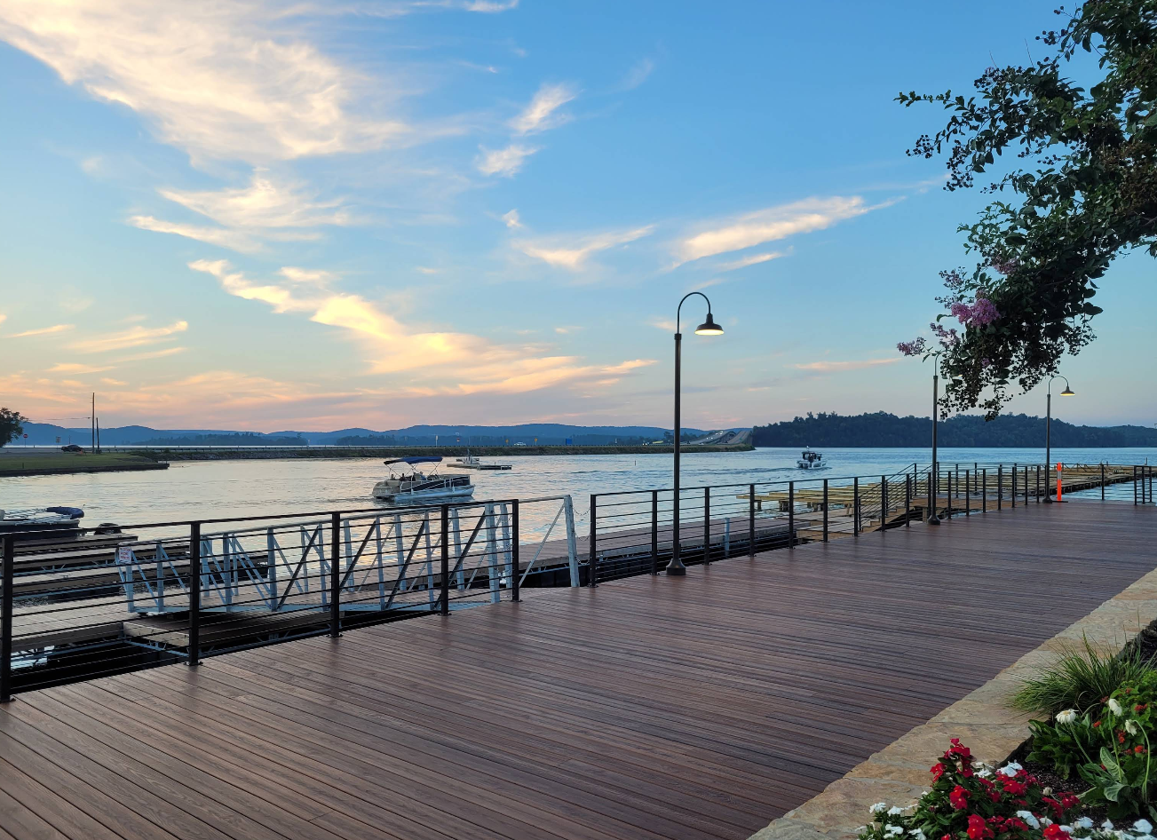 A wooden boardwalk runs alongside a river at sunset, with boats docked on the water—a scene reminiscent of picturesque lake towns. Soft clouds fill the sky, street lamps line the path, and flowers and leaves frame the peaceful setting.