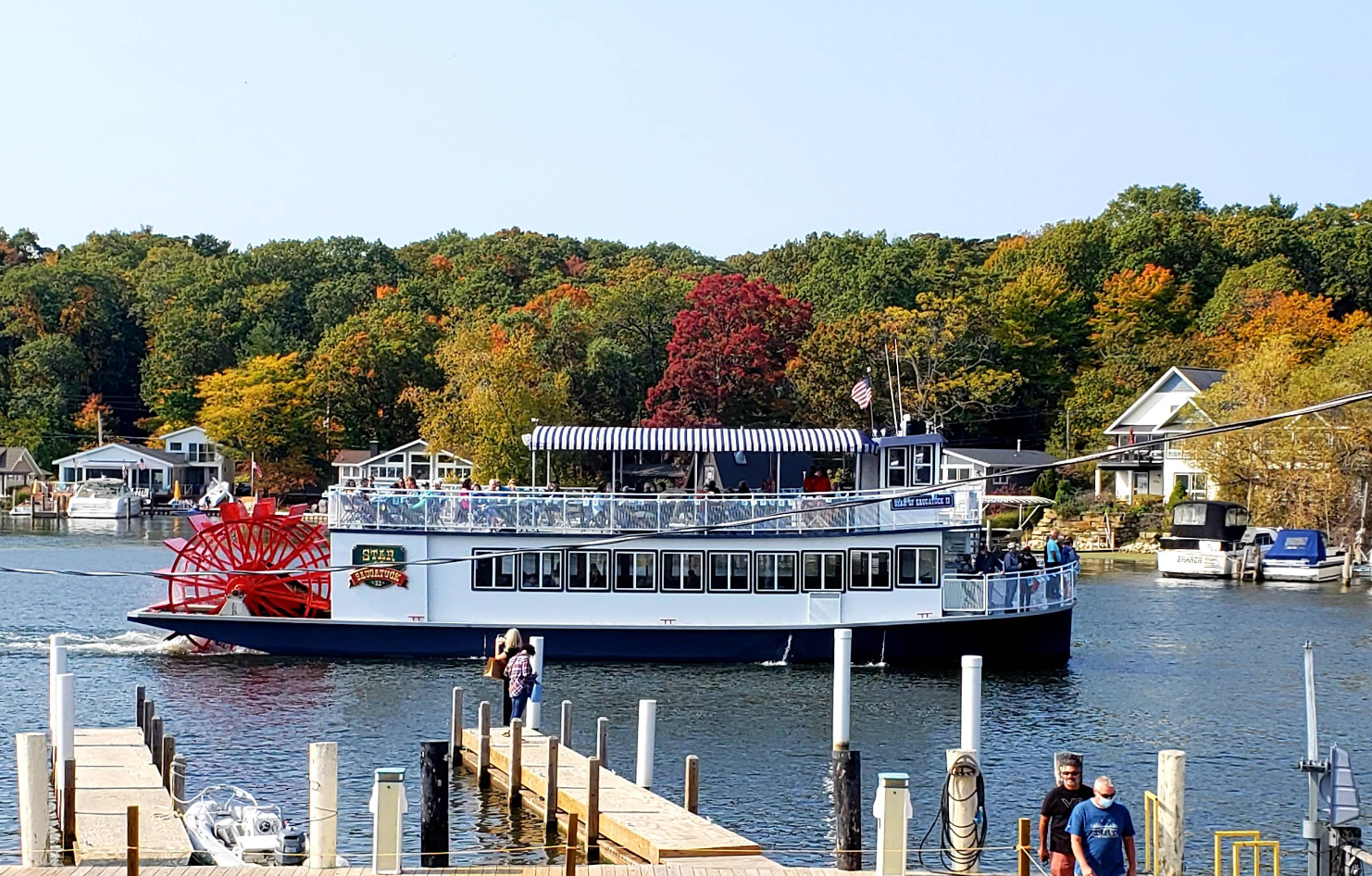 A riverboat with a red paddlewheel sails past docks and waterfront houses in charming lake towns, surrounded by trees with green and autumn-colored leaves under a clear sky. People are visible on the boat and along the dock.
