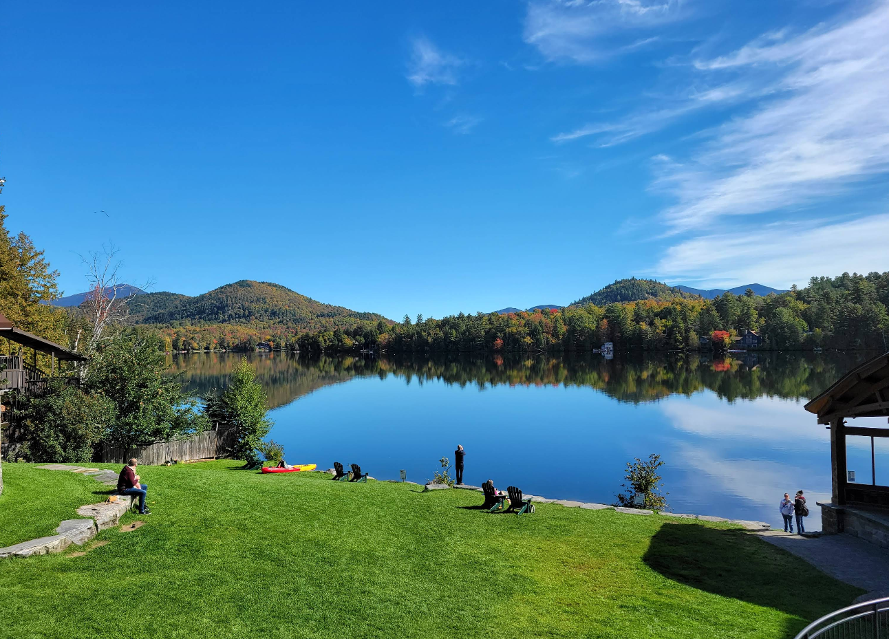 People relax on a green lawn by a calm lake in one of the scenic lake towns, surrounded by forested hills under a clear blue sky, with reflections of trees and mountains shimmering in the water.