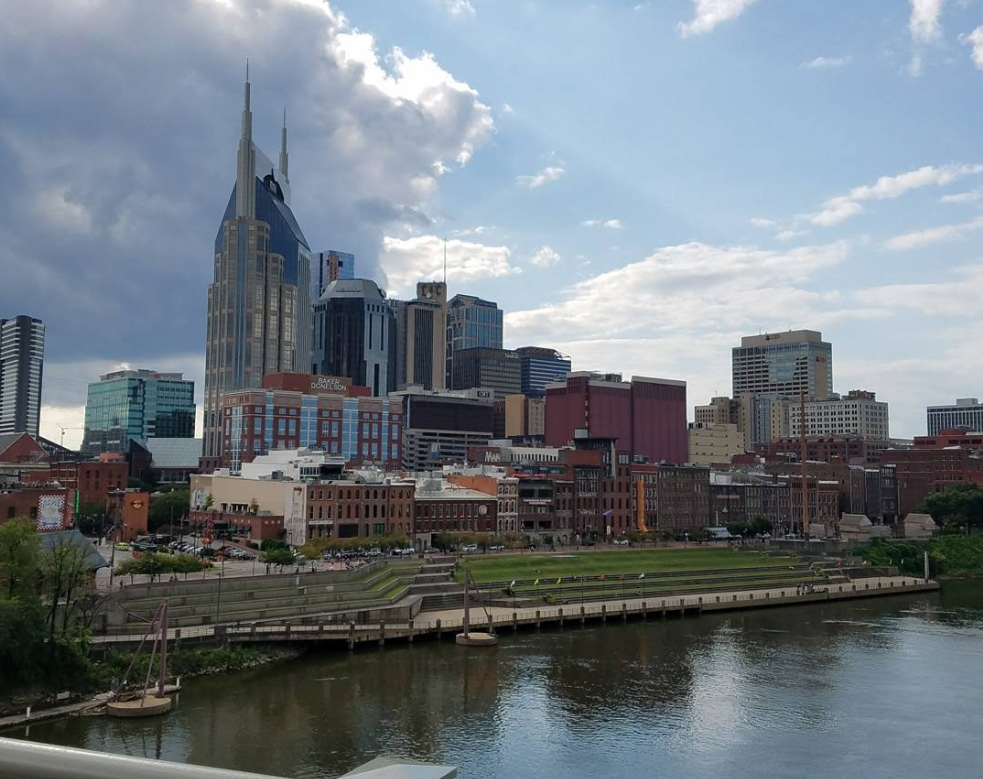 Downtown Nashville skyline with the AT&T “Batman Building” and other high-rises rising behind historic brick buildings, plus a river and grassy foreground—an inviting scene where travelers save while soaking in city charm under a partly cloudy sky.