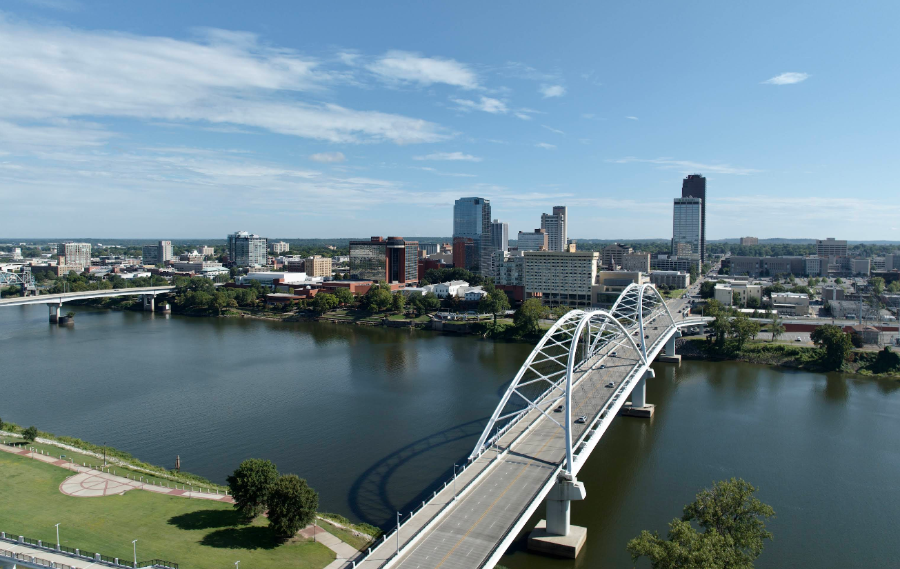 A large arched bridge spans a wide river, leading into a city with tall buildings and a clear blue sky. A park with trees and pathways lines the riverbank, offering travelers save moments to relax before exploring the vibrant city beyond.