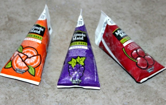 Three triangular Minute Maid juice bar packages on a countertop: orange (orange flavor), purple (grape flavor), and red (cherry flavor). Each package features corresponding fruit images and frosty surfaces.