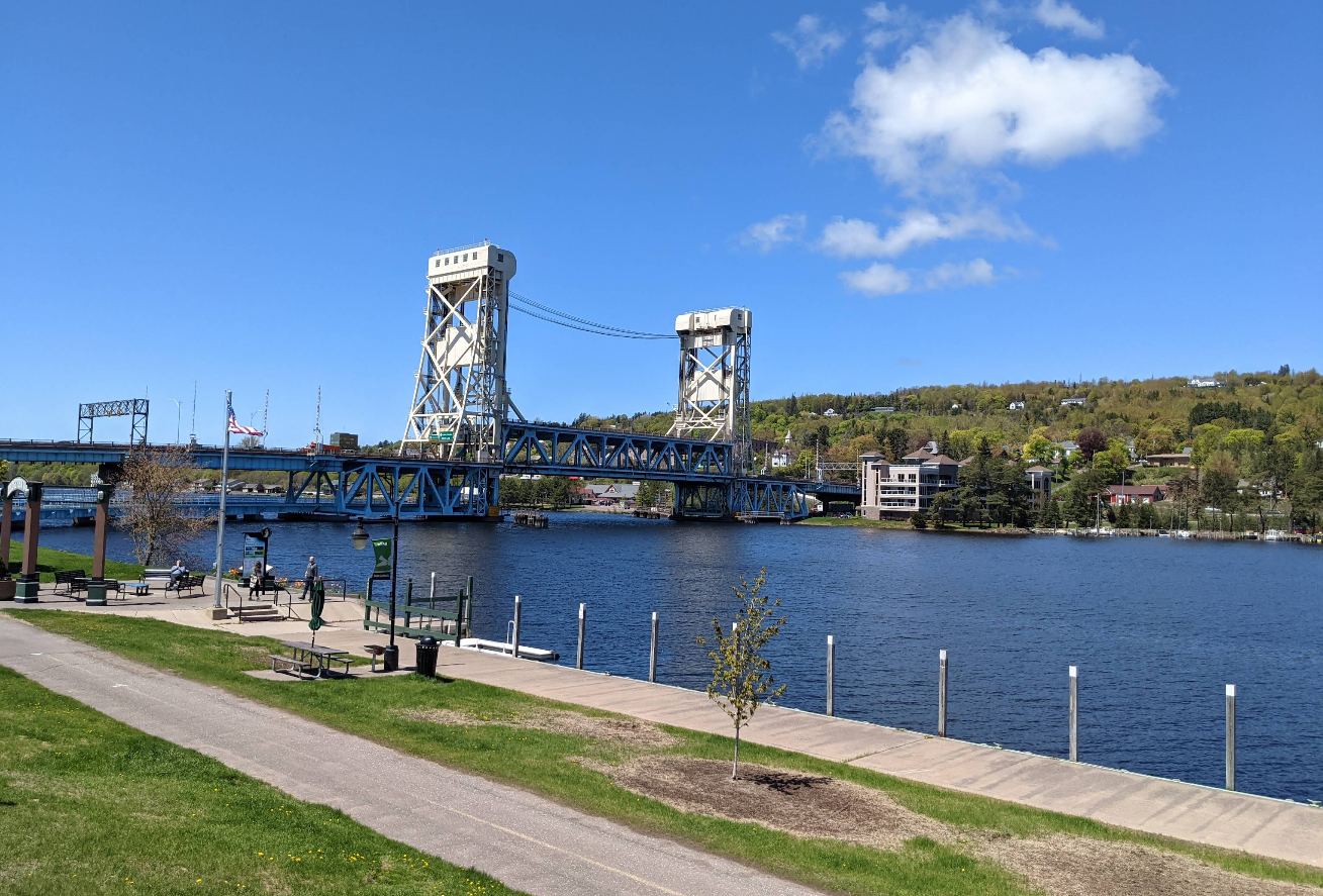 A blue lift bridge spans a river under a clear sky with a few clouds. Travelers save memories as they walk and bike along the riverside path bordered by grass and trees, with hills and buildings in the background.