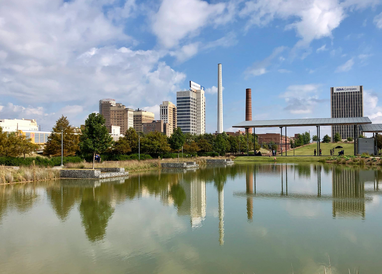 A cityscape with several tall buildings and smokestacks is reflected in a calm pond, surrounded by green grass and trees under a partly cloudy blue sky—an inviting view where travelers save memories of urban tranquility.