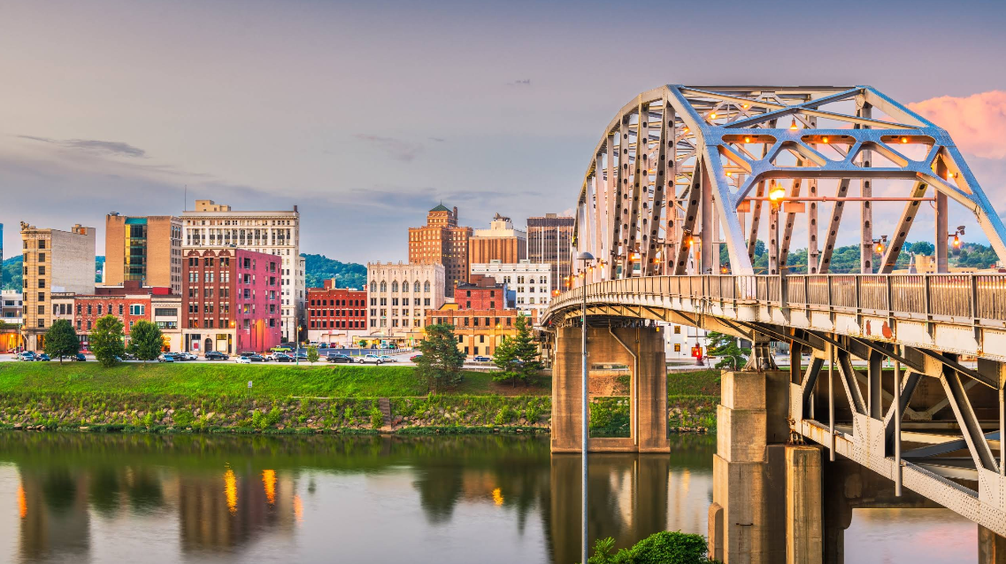 A steel arch bridge crosses a calm river toward a cityscape with colorful historic buildings and modern structures under a soft, early evening sky—a scenic route where savvy travelers save on memorable journeys.