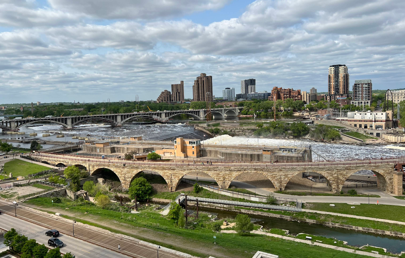 A cityscape featuring a stone arched bridge crossing a river with rapids, a modern skyline in the background, and partly cloudy skies above. Green spaces and roads invite travelers to save time while exploring the vibrant city.