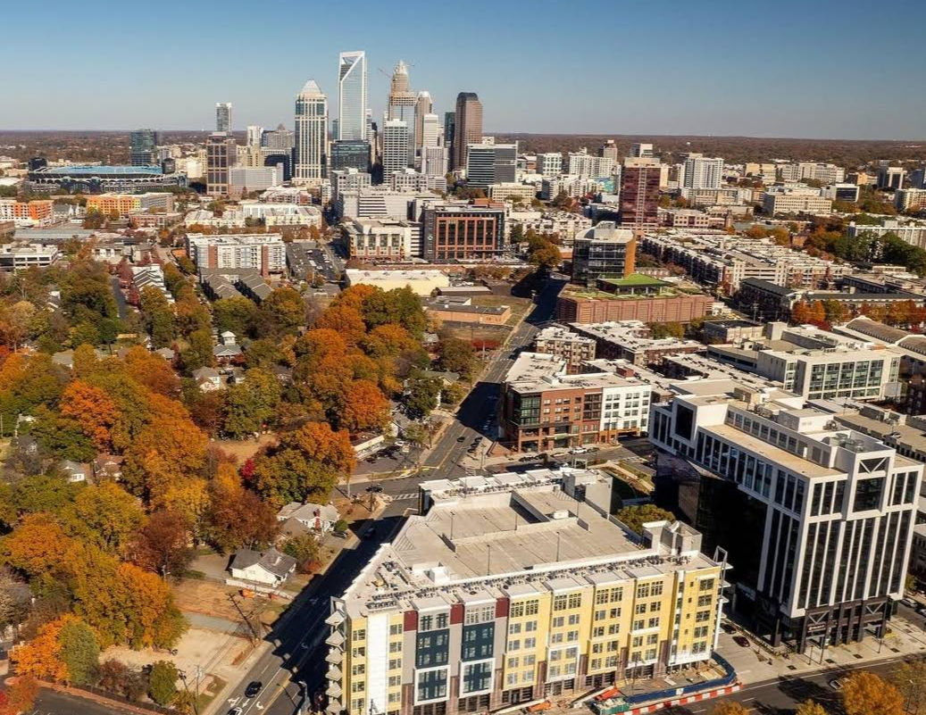 Aerial view of a city with tall skyscrapers in the background and residential buildings surrounded by trees with autumn foliage in the foreground under a clear blue sky—an ideal scene where travelers save memories and capture breathtaking sights.