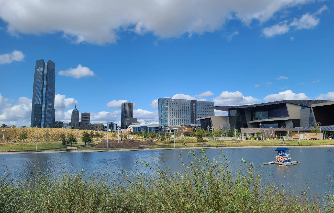 A cityscape with modern high-rise buildings under a blue sky; in the foreground, travelers save on their journey as a small pedal boat with people floats on a calm pond, and greenery lines the water’s edge.