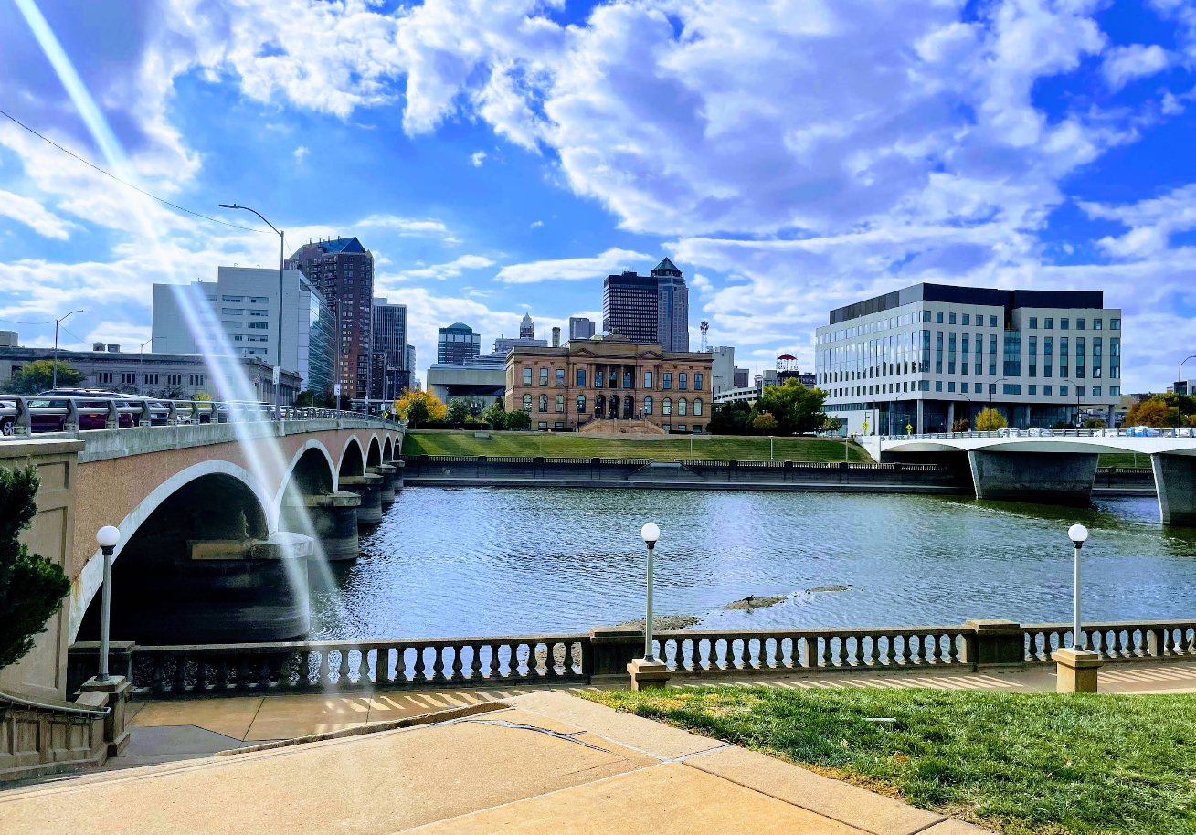 A cityscape with a river running through it, bordered by bridges and modern buildings under a partly cloudy sky. Sunlight creates a bright streak across the scene, beckoning travelers to save moments along grassy and paved walkways in the foreground.