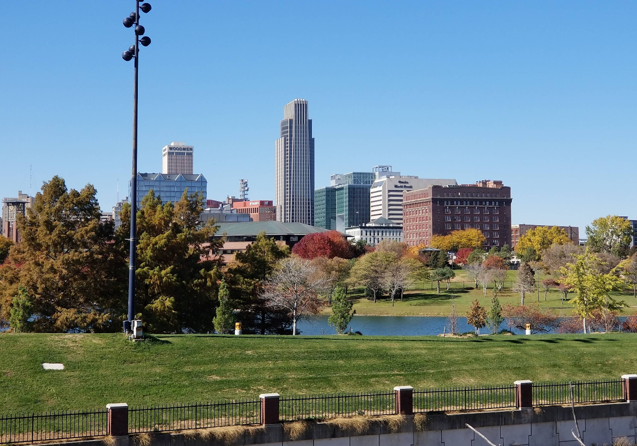 A city skyline with tall buildings rises behind a park lined with autumn-colored trees, next to a small body of water under a clear blue sky—a perfect spot where travelers save memories of peaceful moments.