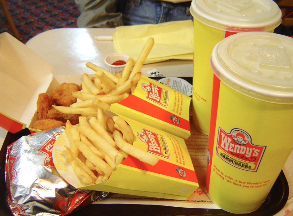 A fast food meal from Wendy’s on a tray, featuring two large cups, two servings of fries, chicken nuggets, a wrapped burger, and a small cup of ketchup.