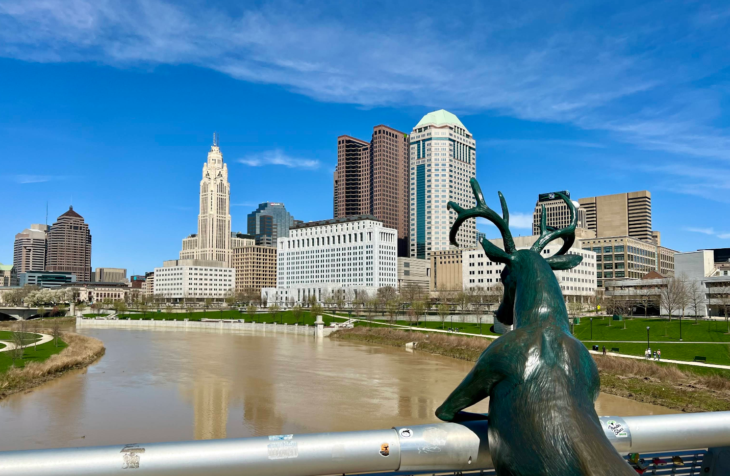 A bronze deer sculpture overlooks a river with the Columbus, Ohio skyline in the background under a clear blue sky. The city's buildings reflect sunlight as travelers save memories strolling along green spaces on the riverbank.