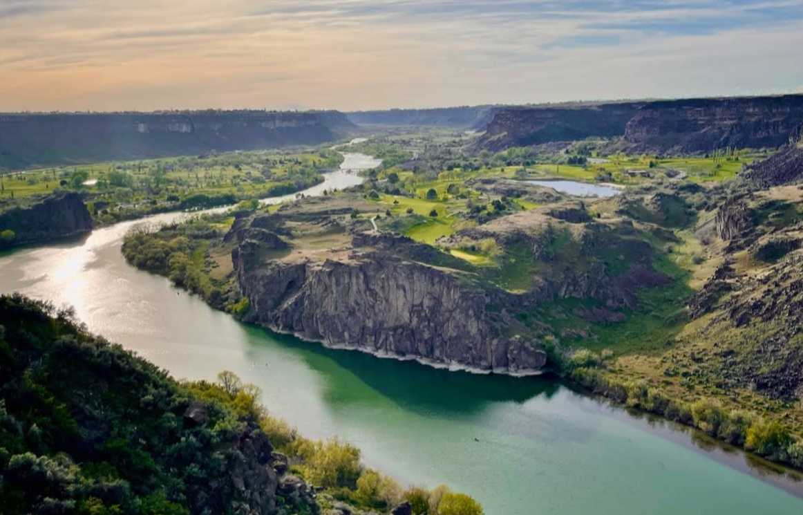 A wide river winds through a lush, green valley surrounded by rocky cliffs under a partly cloudy sky, where sunlight casts a gentle glow over the landscape—a hidden gem where savvy travelers save while enjoying breathtaking views.