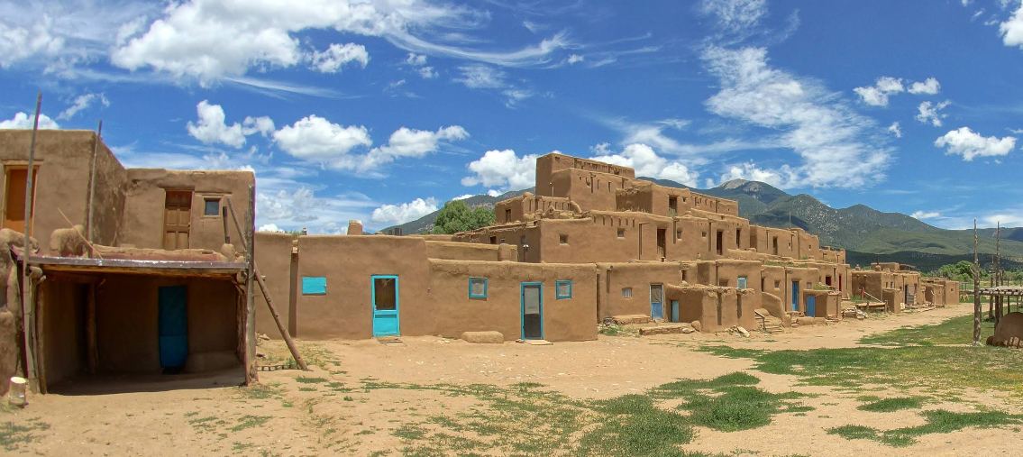 Multi-story adobe buildings with blue doors and windows sit under a bright sky, inviting travelers to save memories as they explore distant green mountains and open land in the foreground.