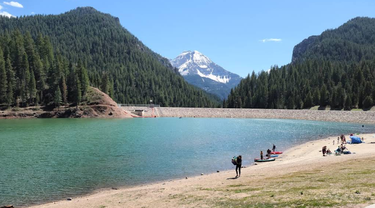 Travelers save while relaxing on a sandy lakeshore bordered by pine forests, with a turquoise lake, dam, and snow-capped mountain in the background under a clear blue sky.