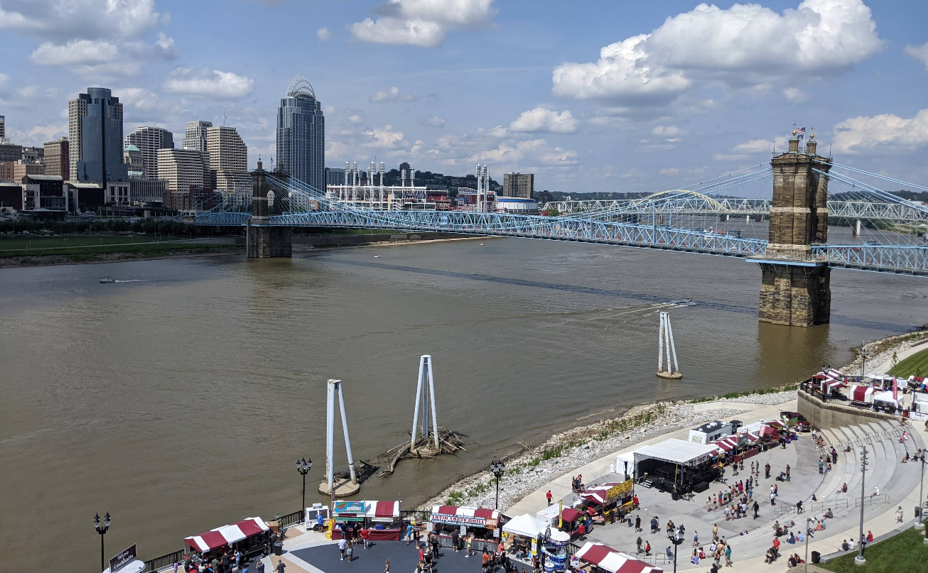 View of a large river with a blue suspension bridge, city skyline in the background, and a lively festival with tents and crowds—travelers save while enjoying the vibrant riverside scene under a partly cloudy sky.