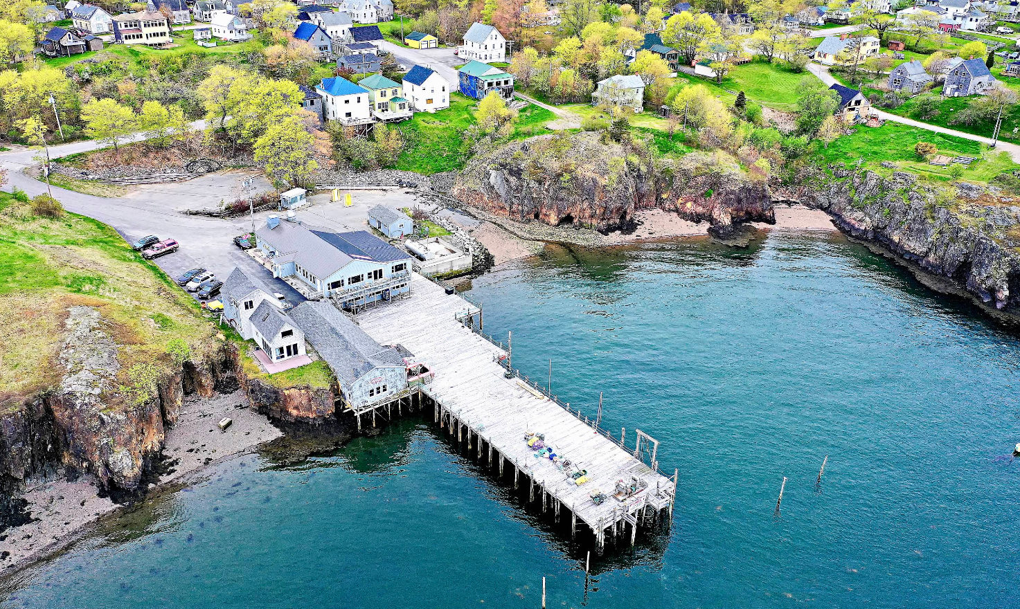 Aerial view of a coastal village with a wooden pier extending into the water, surrounded by rocky cliffs, green trees, and scattered houses on grassy hills—an ideal spot where travelers save while enjoying stunning scenery.