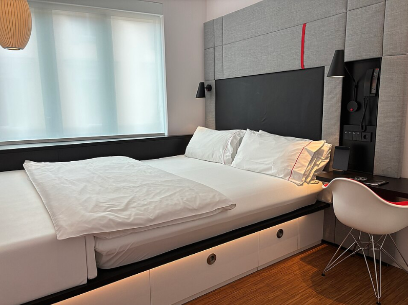 A modern hotel room from leading hotel brands, featuring a neatly made double bed, white linens, a padded headboard, wall-mounted lamps, a desk with a chair, and a large window with sheer curtains letting in soft light.