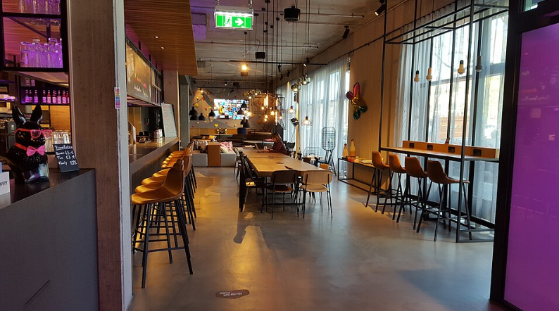 Modern, brightly lit café with high stools at a bar, wooden tables and chairs, large windows with sheer curtains, and contemporary decor. Neon lights and hanging bulbs evoke the inviting vibe of stylish hotel brands.