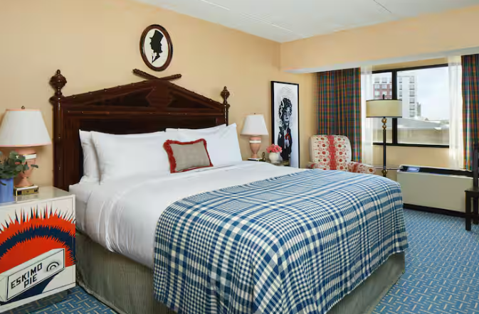 A hotel room from top hotel brands featuring a large bed with a blue plaid blanket, white pillows, red accent pillow, two unique lamps on nightstands, patterned armchair, city-view window, and eclectic framed art on the walls.