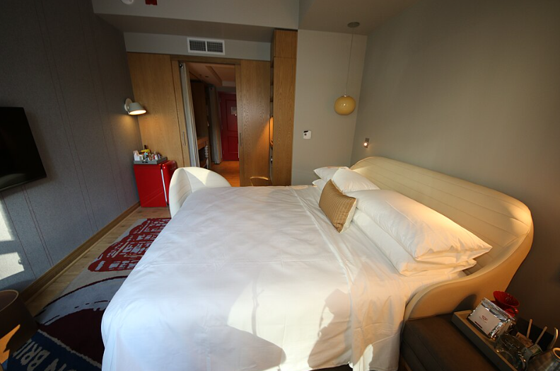 A modern hotel room from leading hotel brands features a neatly made double bed with a cream headboard, white linens, and a beige pillow. Sunlight streams through the window, illuminating the bed. The door and hallway are visible in the background.