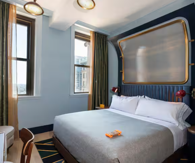 A modern hotel room by leading hotel brands with a large bed, gray bedding, two red lamps, green curtains, tall windows with city views, and a unique gold-framed headboard on a blue accent wall.