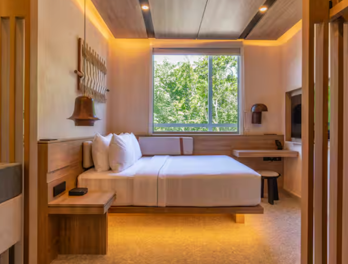 A modern, minimalist hotel room from leading hotel brands, featuring a large bed with white bedding, wooden furniture, soft lighting, and a window overlooking green trees outside.