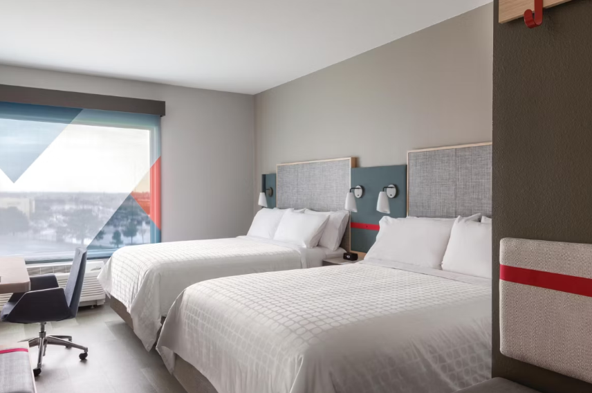 A modern hotel room from top hotel brands featuring two double beds, crisp white bedding, a large window with a view, wall-mounted TV, work desk and chair, and minimalist decor in neutral tones.
