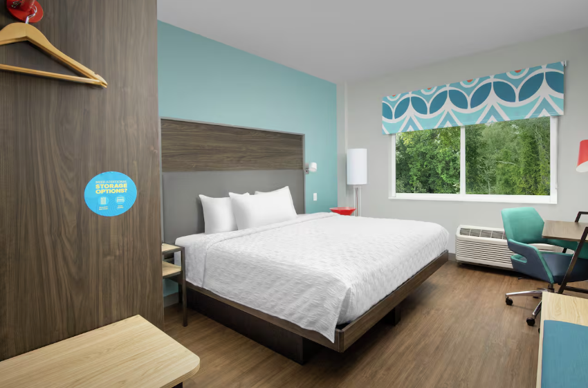 A modern hotel room by top hotel brands, featuring a queen bed, teal accent wall, patterned window shade, large window with greenery views, desk and chair, air conditioner, wall hooks, and wood flooring.