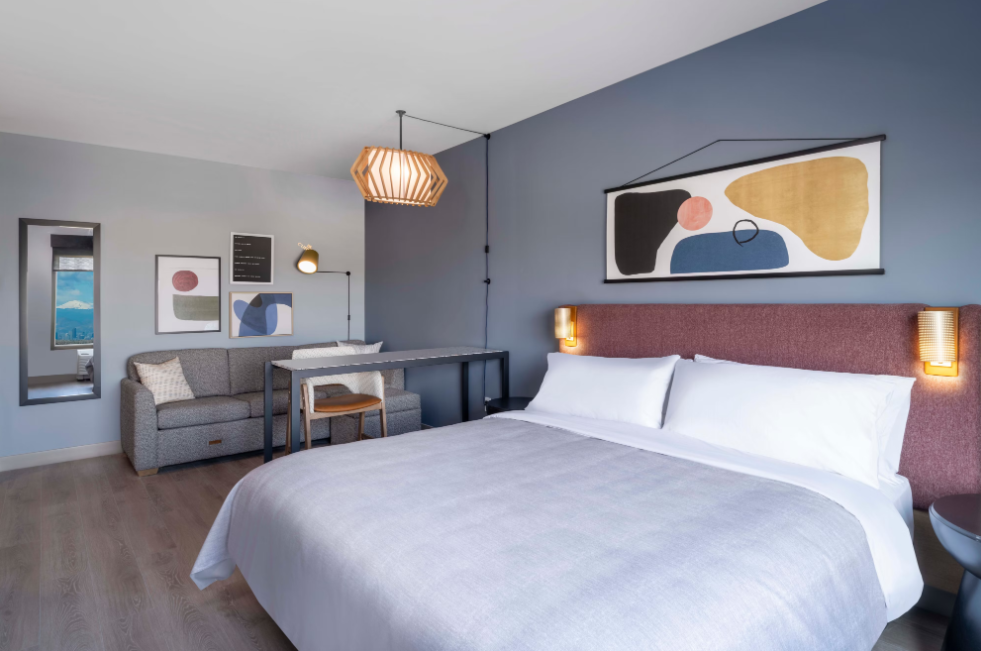 A modern hotel room by leading hotel brands featuring a large bed, abstract wall art, a gray sofa, desk and chair, wooden flooring, and minimalistic lighting set against stylish blue and white walls.