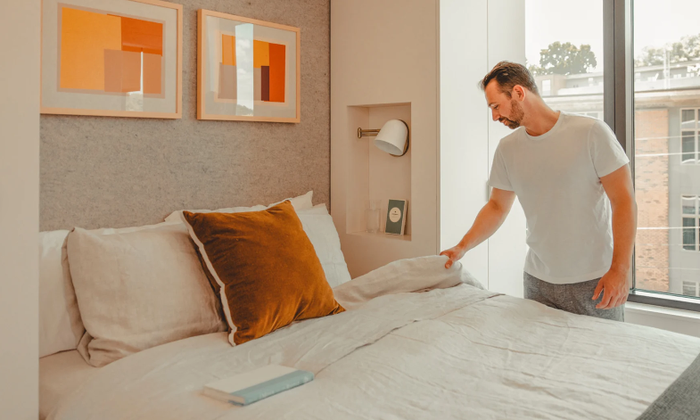 A man in a white t-shirt makes a neatly arranged bed with white bedding and a brown pillow, creating a hotel brands-inspired look in a bright, modern bedroom with large windows and abstract art on the wall.