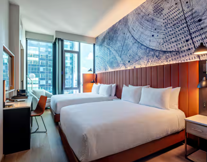 Modern hotel room from leading hotel brands, featuring two large beds with white linens, a blue abstract mural on the wall, a desk with a chair, floor-to-ceiling windows, and city buildings visible outside.