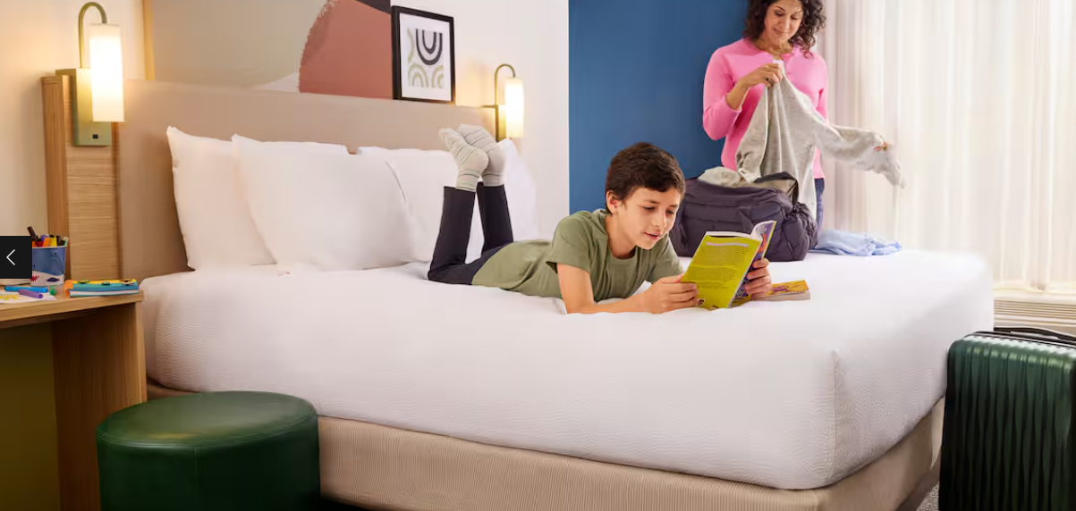 A boy lies on a hotel bed reading a book, while a woman in the background unpacks clothes from a suitcase near the window. The brightly lit, modern room features colorful wall art and decor found in top hotel brands.